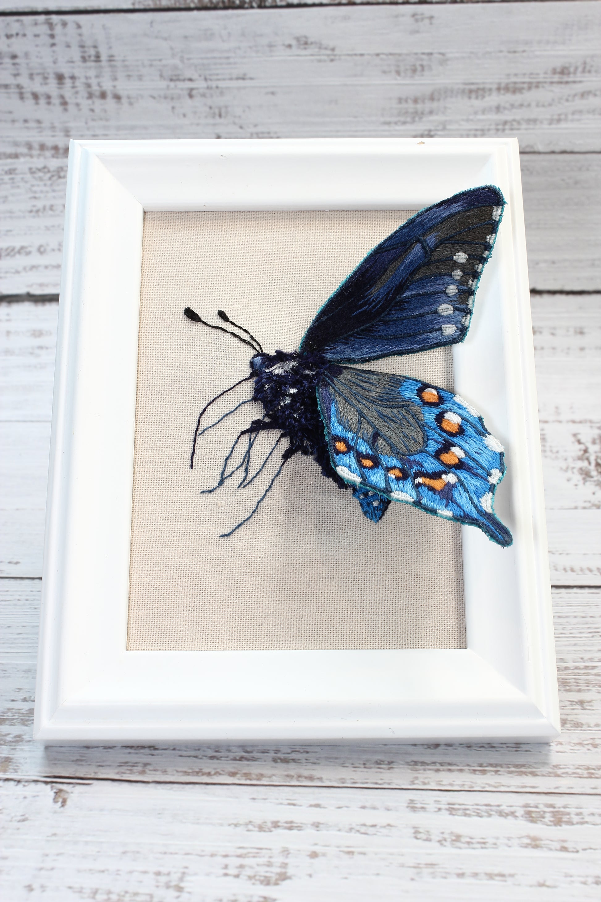 Framed embroidered butterfly with blue and black wings on a wooden surface