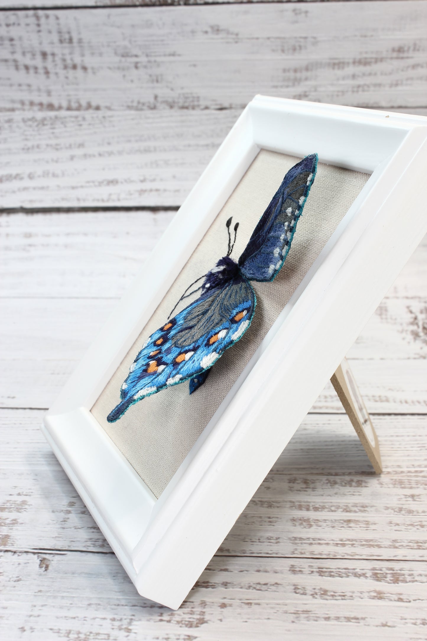 Blue embroidered butterfly  in a white frame on a wooden surface
