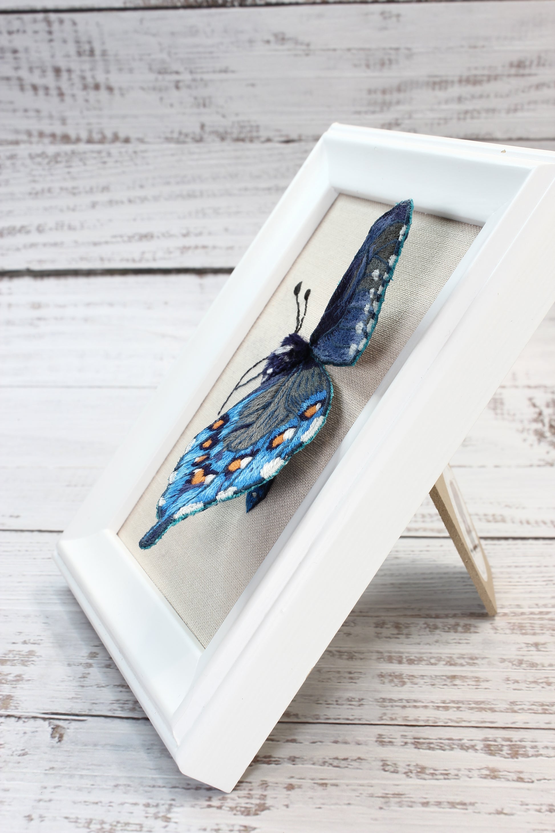 Blue embroidered butterfly  in a white frame on a wooden surface