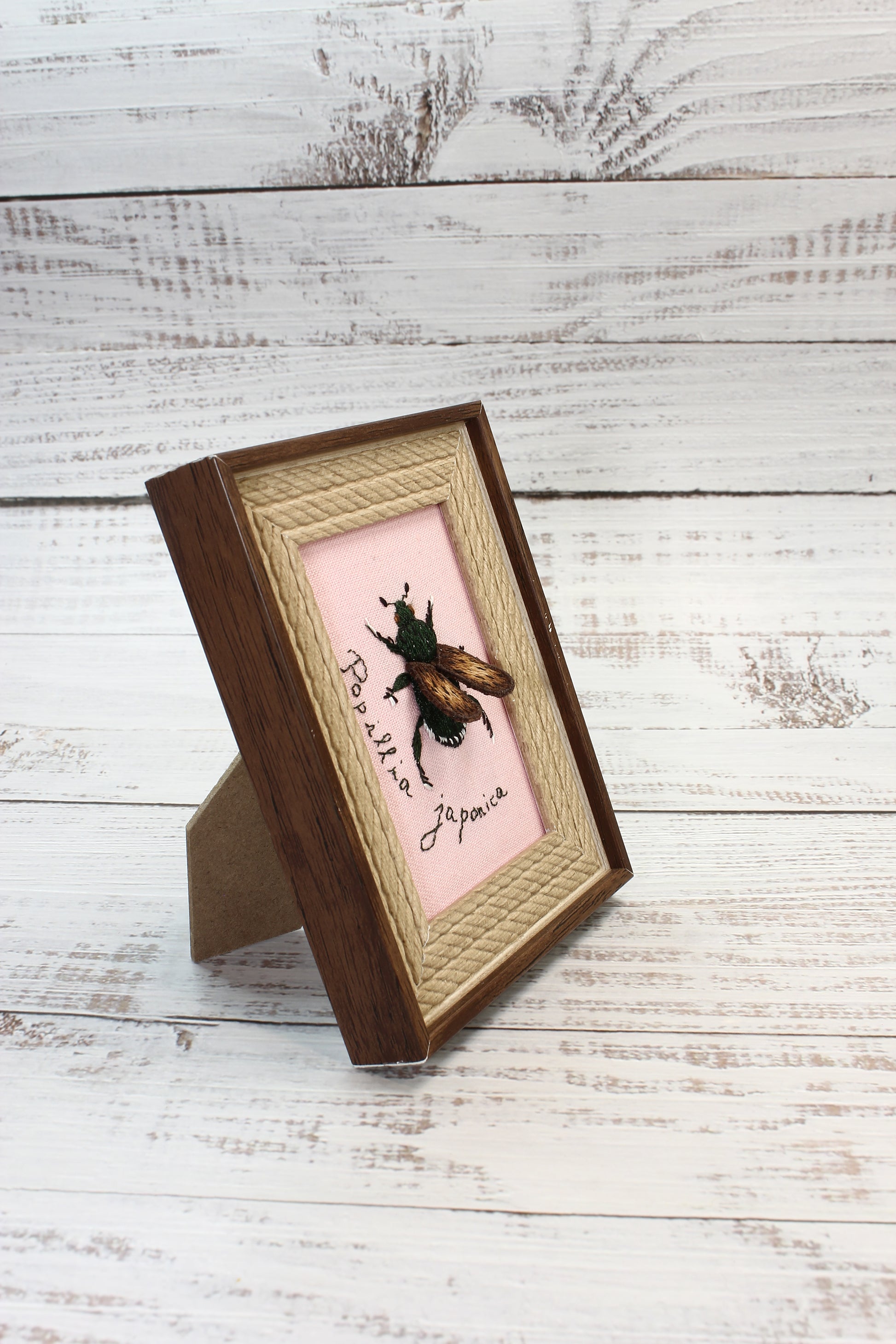 Embroidered insect on a pink background with wooden frame on a light wooden surface